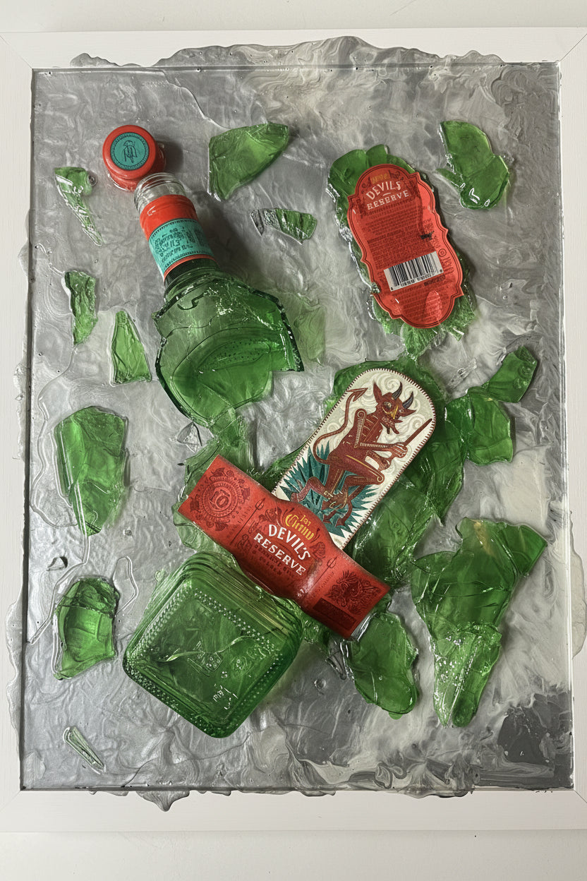 A square decorative artwork featuring a broken green glass bottle and shards arranged on a textured silver background, with a red label reading Devil's Riverside visible on one piece.